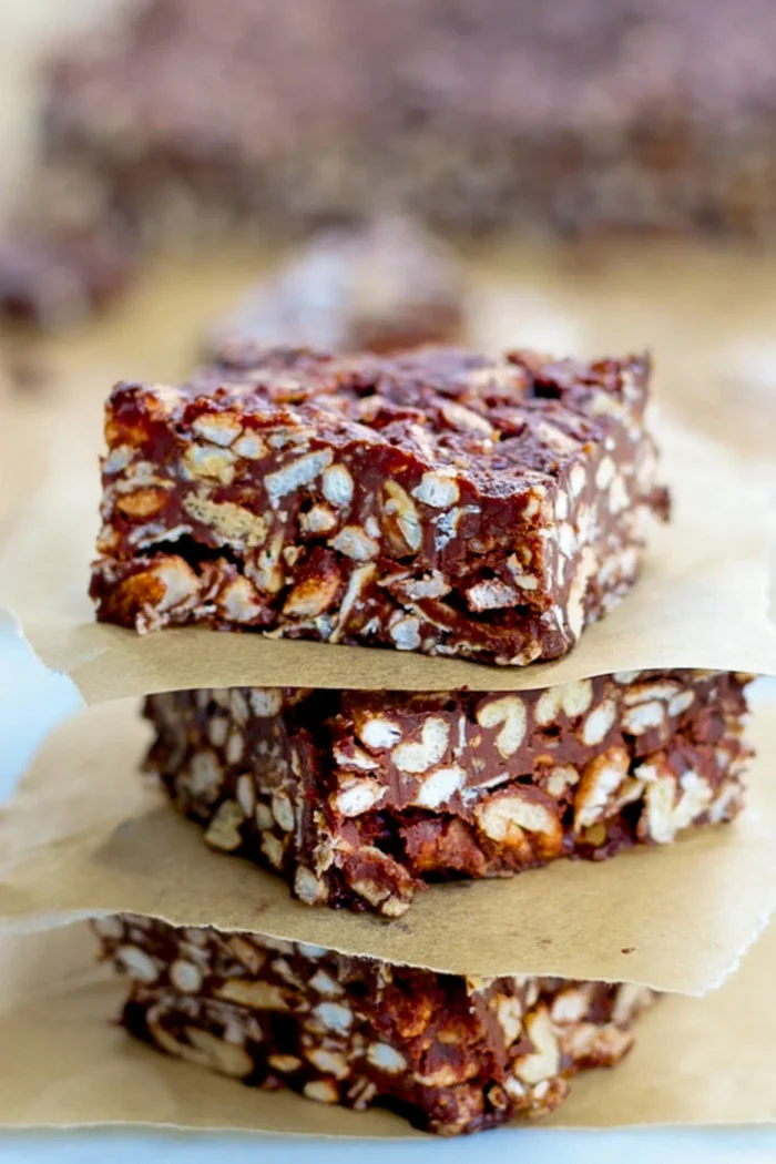 Tres cuadrados de arroz crujiente con chocolate, o barras de chocolate, se apilan sobre papel de pergamino marrón. Rellenas de arroz inflado y recubiertas de chocolate, estas barritas tienen un aspecto texturizado y troceado. El fondo difuminado atrae la atención hacia las tentadoras golosinas.