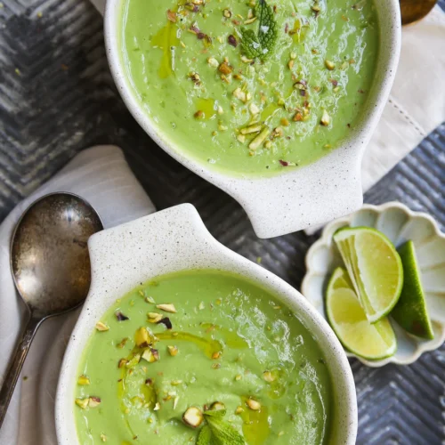 Dos cuencos de cremosa sopa fría de aguacate se adornan con nueces picadas, hojas de menta y un chorrito de aceite. Cerca, sobre una superficie de textura gris, hay un pequeño plato con gajos de lima, una servilleta, una cuchara y utensilios dorados.