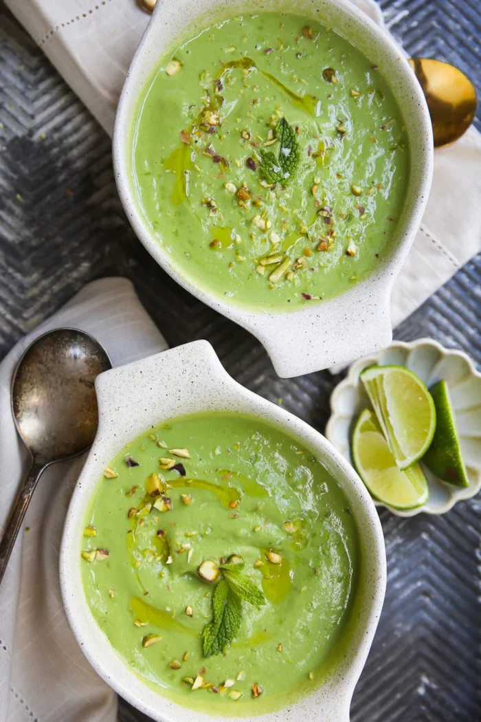 Dos cuencos de cremosa sopa fría de aguacate se adornan con nueces picadas, hojas de menta y un chorrito de aceite. Cerca, sobre una superficie de textura gris, hay un pequeño plato con gajos de lima, una servilleta, una cuchara y utensilios dorados.