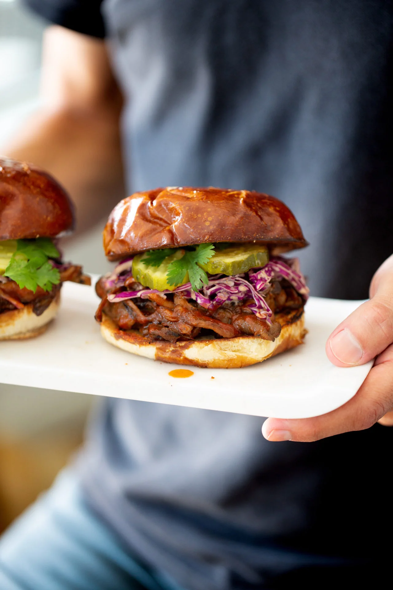 Una persona con camisa oscura sostiene un plato blanco con un sándwich vegano de barbacoa. El sándwich lleva un pan brillante, carne desmenuzada a la barbacoa, ensalada morada, pepinillos y cilantro. Se ve parcialmente otro sándwich similar en el plato.