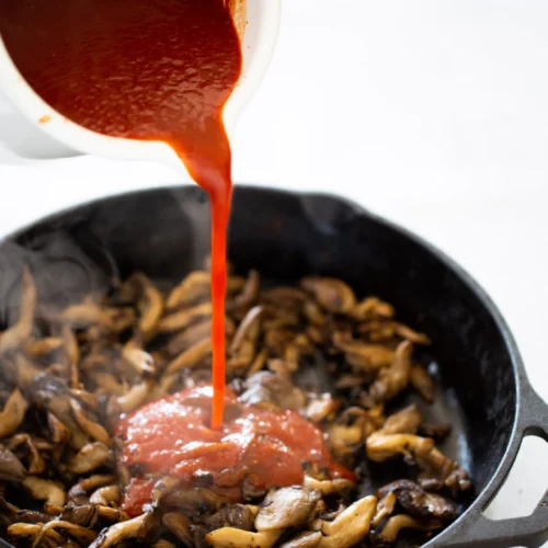 Primer plano de una sartén de hierro fundido negra llena de champiñones salteados, mientras una mano vierte salsa de tomate roja brillante desde un recipiente blanco. El vapor que sale de los champiñones calientes realza el proceso de cocción.
