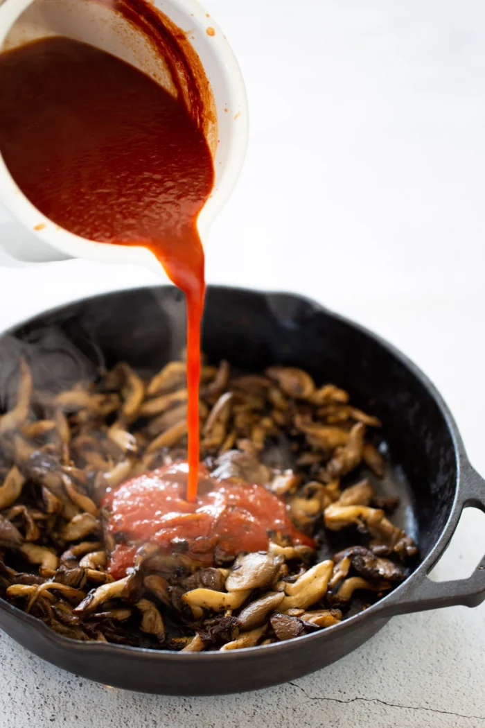 Primer plano de una sartén de hierro fundido negra llena de champiñones salteados, mientras una mano vierte salsa de tomate roja brillante desde un recipiente blanco. El vapor que sale de los champiñones calientes realza el proceso de cocción.