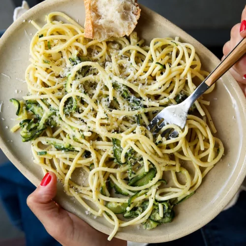 Una persona sostiene un plato ovalado de espaguetis mezclados con calabacín, con queso rallado y hierbas aromáticas. Un tenedor hace girar la pasta, y en el plato hay un trozo de pan. La persona lleva esmalte de uñas rojo y viste una blusa de encaje y pulseras.