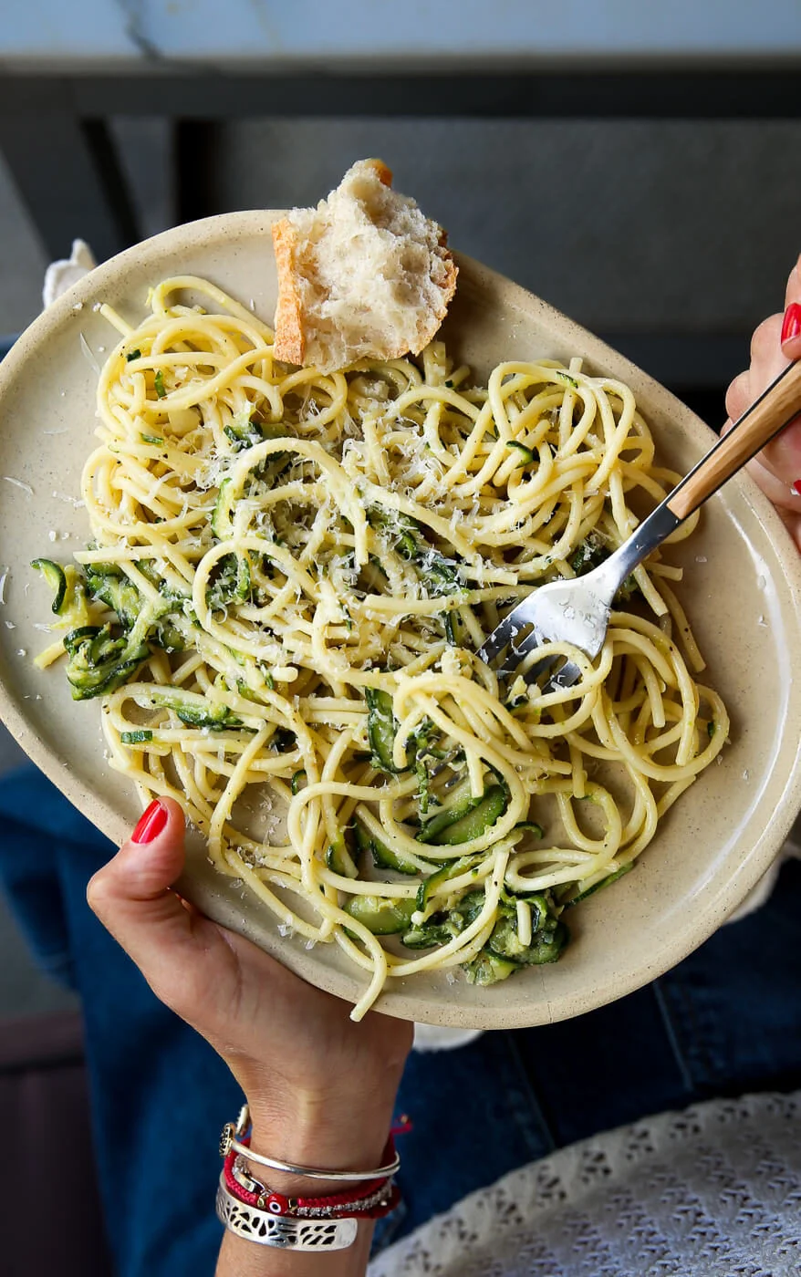 Una persona con esmalte rojo sostiene un plato ovalado beige con Pasta de Calabacita y Parmesano—una receta rápida y fácil de verano—girando un tenedor sobre el espagueti con queso, mientras un pan crujiente descansa al borde del plato. Lleva una blusa blanca de encaje, jeans y pulseras.