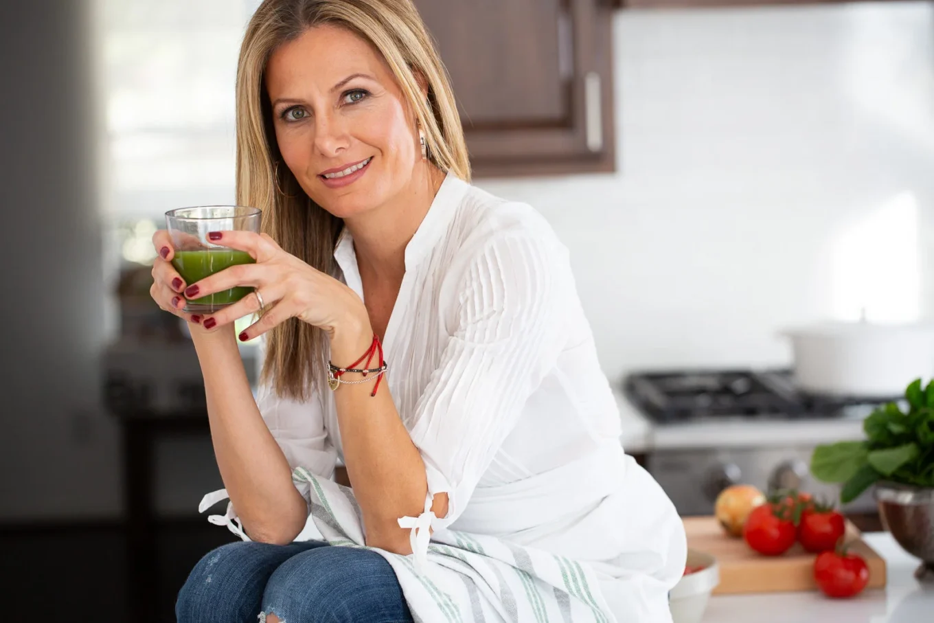 Una mujer de pelo largo y rubio, con camisa blanca y vaqueros azules, está sentada en una cocina con un vaso de zumo verde en la mano. Sonríe a la cámara; en la encimera del fondo se ven verduras frescas y una olla blanca.