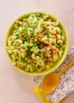 Un cuenco amarillo lleno de pasta de macarrones mezclada con guisantes verdes, zanahorias cortadas en dados, garbanzos y guindillas verdes cortadas en rodajas. El cuenco está sobre una superficie clara junto a una cuchara amarilla que descansa sobre una servilleta blanca con dibujos geométricos negros.