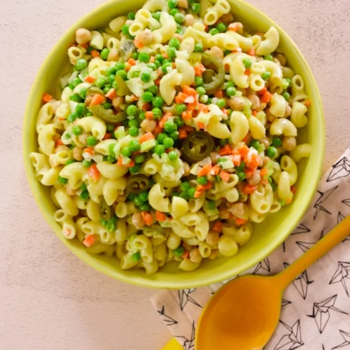Un cuenco amarillo lleno de pasta de macarrones mezclada con guisantes verdes, zanahorias cortadas en dados, garbanzos y guindillas verdes cortadas en rodajas. El cuenco está sobre una superficie clara junto a una cuchara amarilla que descansa sobre una servilleta blanca con dibujos geométricos negros.