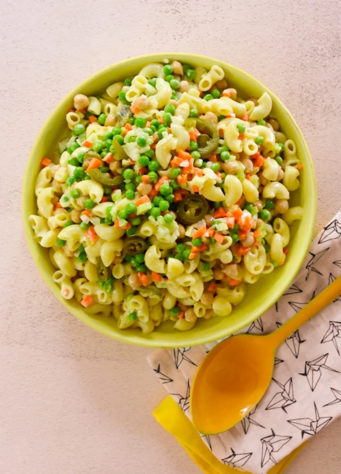 Un cuenco amarillo lleno de pasta de macarrones mezclada con guisantes verdes, zanahorias cortadas en dados, garbanzos y guindillas verdes cortadas en rodajas. El cuenco está sobre una superficie clara junto a una cuchara amarilla que descansa sobre una servilleta blanca con dibujos geométricos negros.