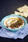 Sobre un mantel a rayas, un cuenco de cremoso hummus blanco con cebolla caramelizada, hierbas verdes y un poco de pimentón. El cuenco verde azulado con motivos blancos se combina con trozos de pan de pita ligeramente desenfocados al fondo.