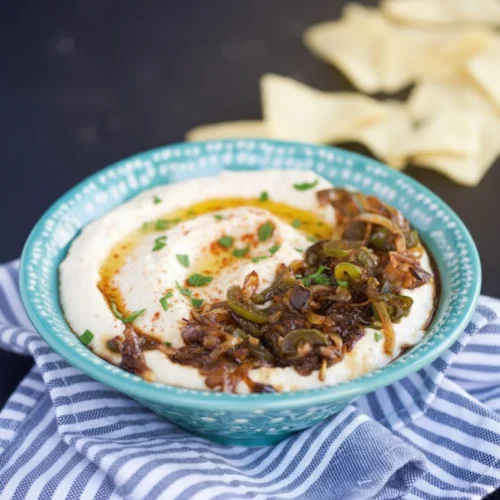 Sobre un mantel a rayas, un cuenco de cremoso hummus blanco con cebolla caramelizada, hierbas verdes y un poco de pimentón. El cuenco verde azulado con motivos blancos se combina con trozos de pan de pita ligeramente desenfocados al fondo.