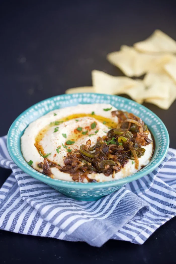 Sobre un mantel a rayas, un cuenco de cremoso hummus blanco con cebolla caramelizada, hierbas verdes y un poco de pimentón. El cuenco verde azulado con motivos blancos se combina con trozos de pan de pita ligeramente desenfocados al fondo.