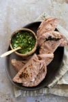 En un plato ovalado oscuro hay cinco tortillas de maíz azul dobladas y ligeramente chamuscadas, rellenas de queso. Junto a ellas hay un cuenco pequeño de madera con salsa verde con trozos de cebolla picada y una cuchara de madera. El plato descansa sobre una servilleta beige, colocada a su vez sobre una superficie de piedra.