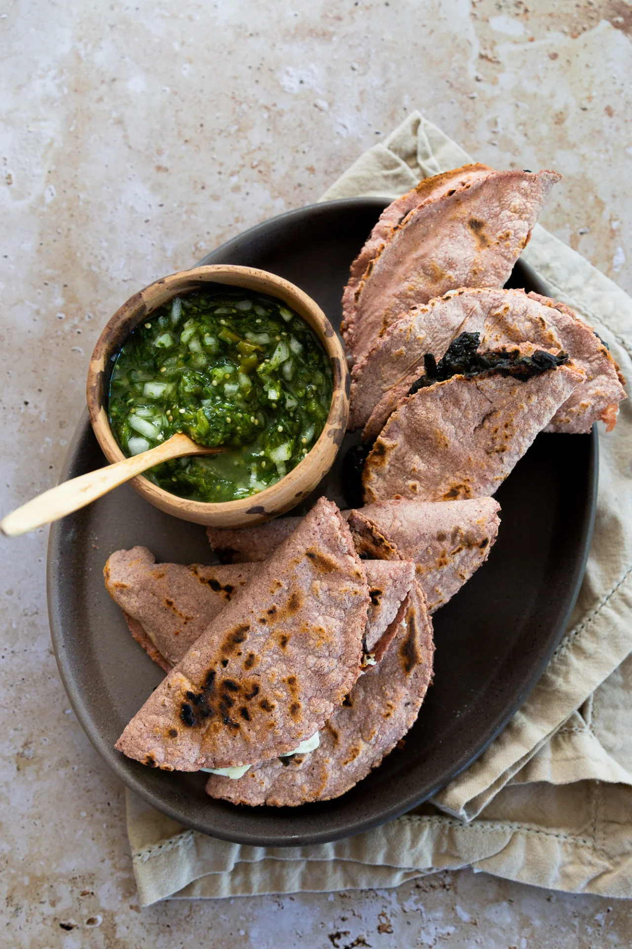 En un plato ovalado oscuro hay cinco tortillas de maíz azul dobladas y ligeramente chamuscadas, rellenas de queso. Junto a ellas hay un cuenco pequeño de madera con salsa verde con trozos de cebolla picada y una cuchara de madera. El plato descansa sobre una servilleta beige, colocada a su vez sobre una superficie de piedra.