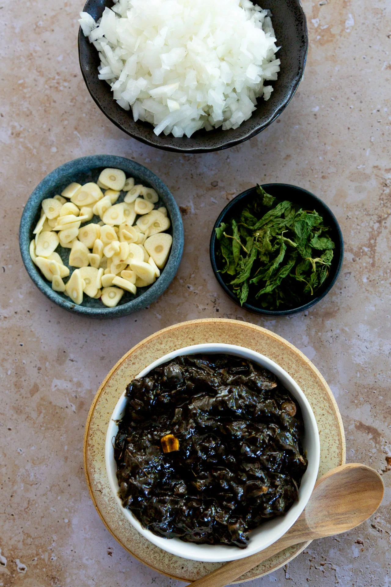Cuatro tazones sobre una superficie de piedra clara con cebolla blanca picada, ajo en rodajas, epazote fresco y huitlacoche, los ingredientes para las quesadillas de huitlacoche, con una cuchara de madera al lado.