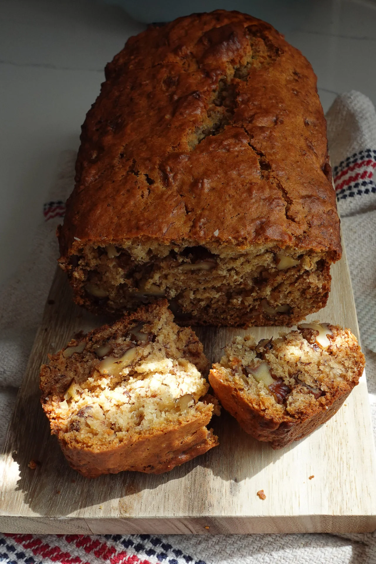 Sobre una tabla de madera hay una hogaza de Pan de plátano con nueces picadas. Se cortan dos rebanadas por delante, revelando un interior húmedo y lleno de nueces. El pan está dorado y descansa sobre un paño de cocina texturizado con rayas rojas y azules.