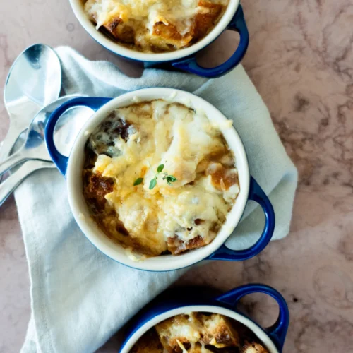 Tres cazuelitas con asas azules rellenas de sopa de cebolla vegana, cubiertas de queso fundido y pan tostado, están dispuestas verticalmente sobre una servilleta de tela de color claro. Dos cucharas metálicas descansan a un lado sobre una superficie de mármol.