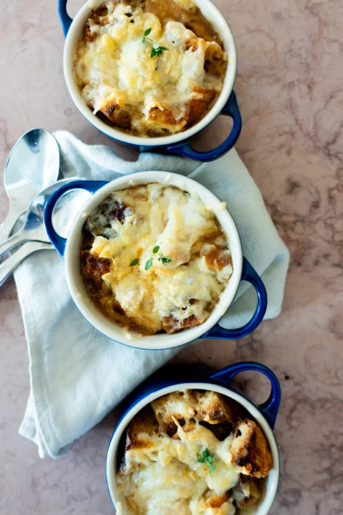 Tres cazuelitas con asas azules rellenas de sopa de cebolla vegana, cubiertas de queso fundido y pan tostado, están dispuestas verticalmente sobre una servilleta de tela de color claro. Dos cucharas metálicas descansan a un lado sobre una superficie de mármol.