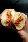 Dos manos sostienen un bocadillo cortado por la mitad, mostrando chorizo mexicano sazonado, rodajas de tomate, cebolla roja, lechuga verde y mayonesa dentro de un bollo blanco suave y redondo. El fondo oscuro hace que el sándwich estilo torta aparezca nítidamente enfocado.