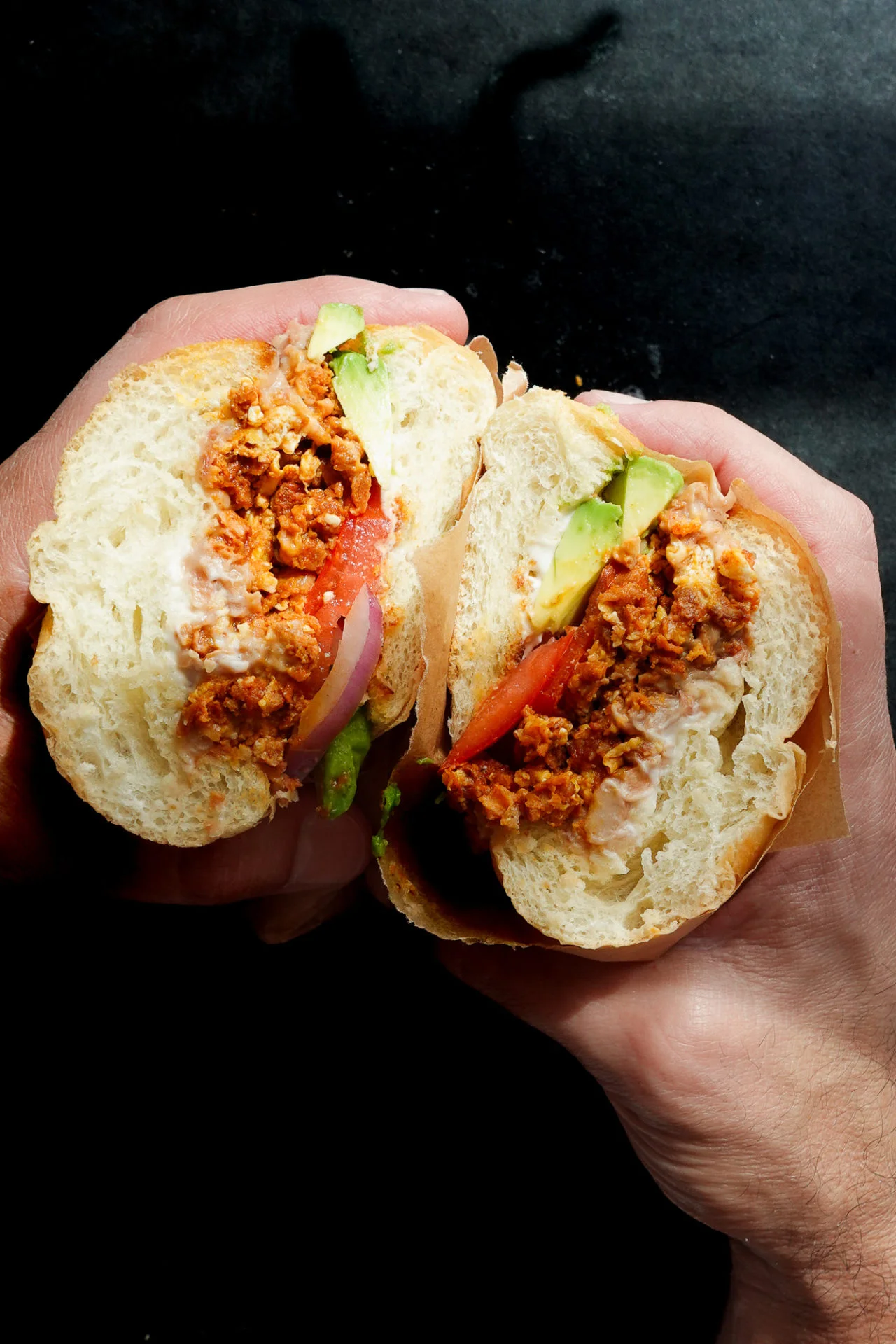Dos manos sostienen un bocadillo cortado por la mitad, mostrando chorizo mexicano sazonado, rodajas de tomate, cebolla roja, lechuga verde y mayonesa dentro de un bollo blanco suave y redondo. El fondo oscuro hace que el sándwich estilo torta aparezca nítidamente enfocado.
