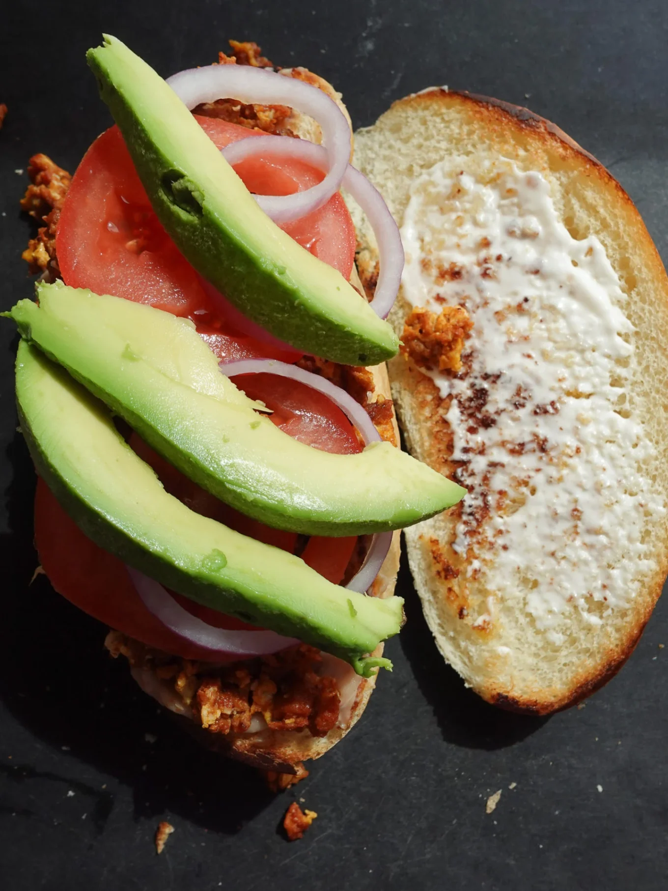 Un sándwich mexicano abierto en pan negro que lleva chorizo cocido desmenuzado, rodajas de tomate, aros de cebolla crudos y aguacate en una mitad, mientras que la otra está untada con mayonesa. Esta Torta de Chorizo descansa sobre una superficie oscura, iluminada desde arriba.