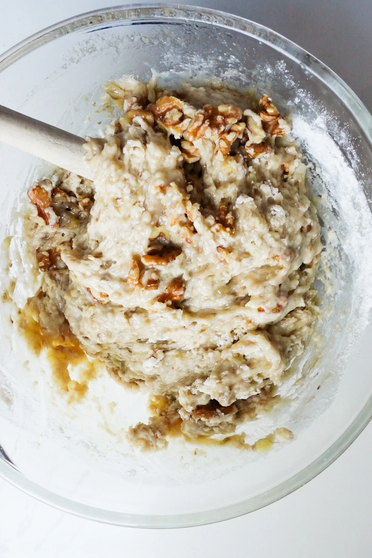 Un cuenco de cristal lleno de masa húmeda de pan de plátano, con trozos visibles de plátano y nueces, y algo de harina sin mezclar en los bordes. Dentro hay una cuchara de madera, lo que indica que la masa se está mezclando a mano en una encimera blanca casi limpia.