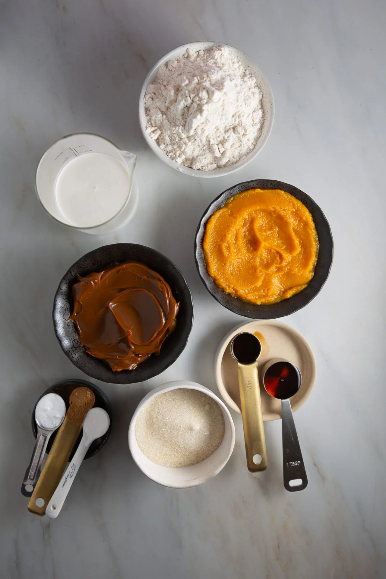 An overhead view of baking ingredients for pumpkin muffins: flour, milk, pumpkin puree, dulce de leche filling, sugar, spices, and more in bowls with measuring spoons or cups, all arranged on a light marble surface.