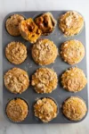 A muffin tin holds twelve pumpkin muffins with streusel topping. One muffin is cut open, revealing a gooey dulce de leche filling inside. Crumbs are scattered on the muffins and tin, set against a light marble countertop.