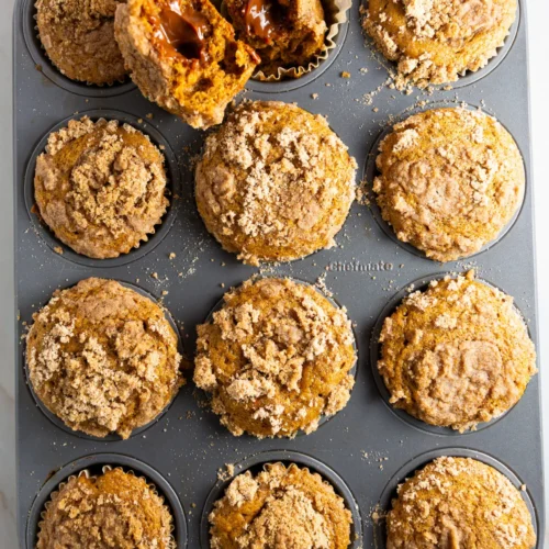 A muffin tin holds twelve pumpkin muffins with streusel topping. One muffin is cut open, revealing a gooey dulce de leche filling inside. Crumbs are scattered on the muffins and tin, set against a light marble countertop.