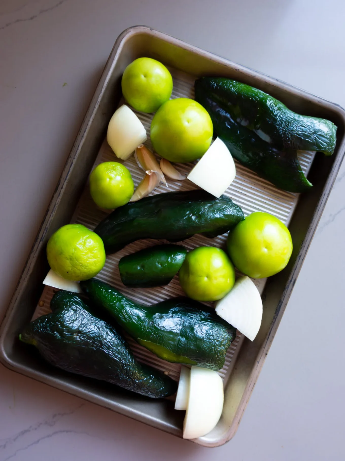 Una bandeja para hornear contiene chiles poblanos verdes crudos, tomatillos, trozos de cebolla blanca, un jalapeño y dientes de ajo, todo listo para ser asado para una sabrosa Salsa Verde de Poblano en una encimera de cocina de color claro.