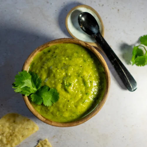 A wooden bowl filled with Salsa Verde garnished with cilantro sits on a light stone surface. Nearby are a silver spoon on a small plate and two tortilla chips. The lighting highlights the salsa’s smooth texture and hints of roasted poblano.