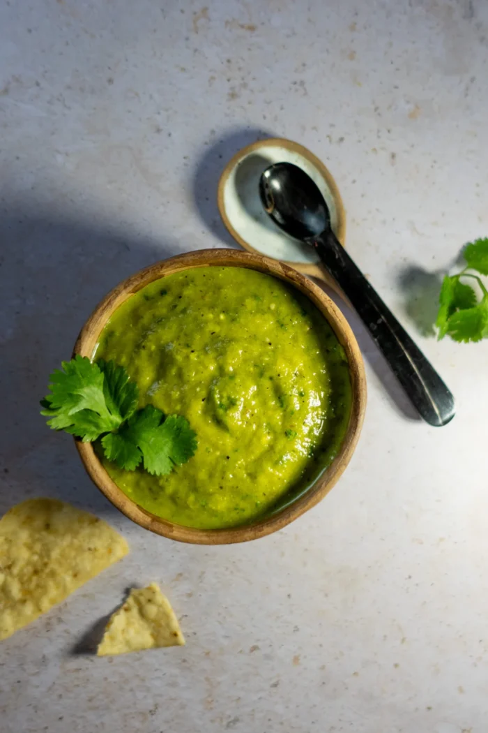 A wooden bowl filled with Salsa Verde garnished with cilantro sits on a light stone surface. Nearby are a silver spoon on a small plate and two tortilla chips. The lighting highlights the salsa’s smooth texture and hints of roasted poblano.