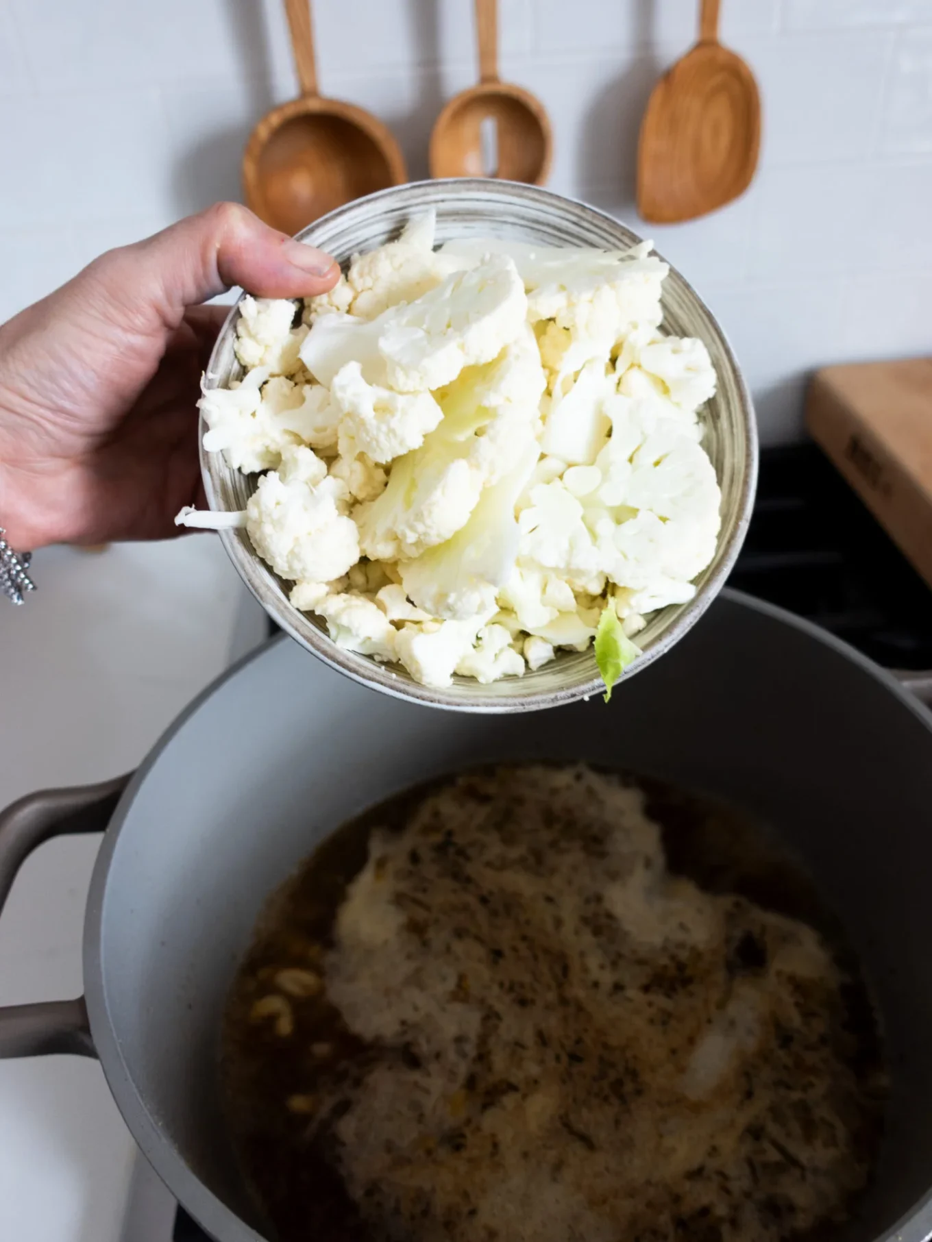 A hand holds a bowl of raw cauliflower florets over a gray pot of simmering broth, ready to create a creamy White Bean Chili. Wooden utensils hang on a white tile wall in the background, adding warmth to the inviting kitchen scene.