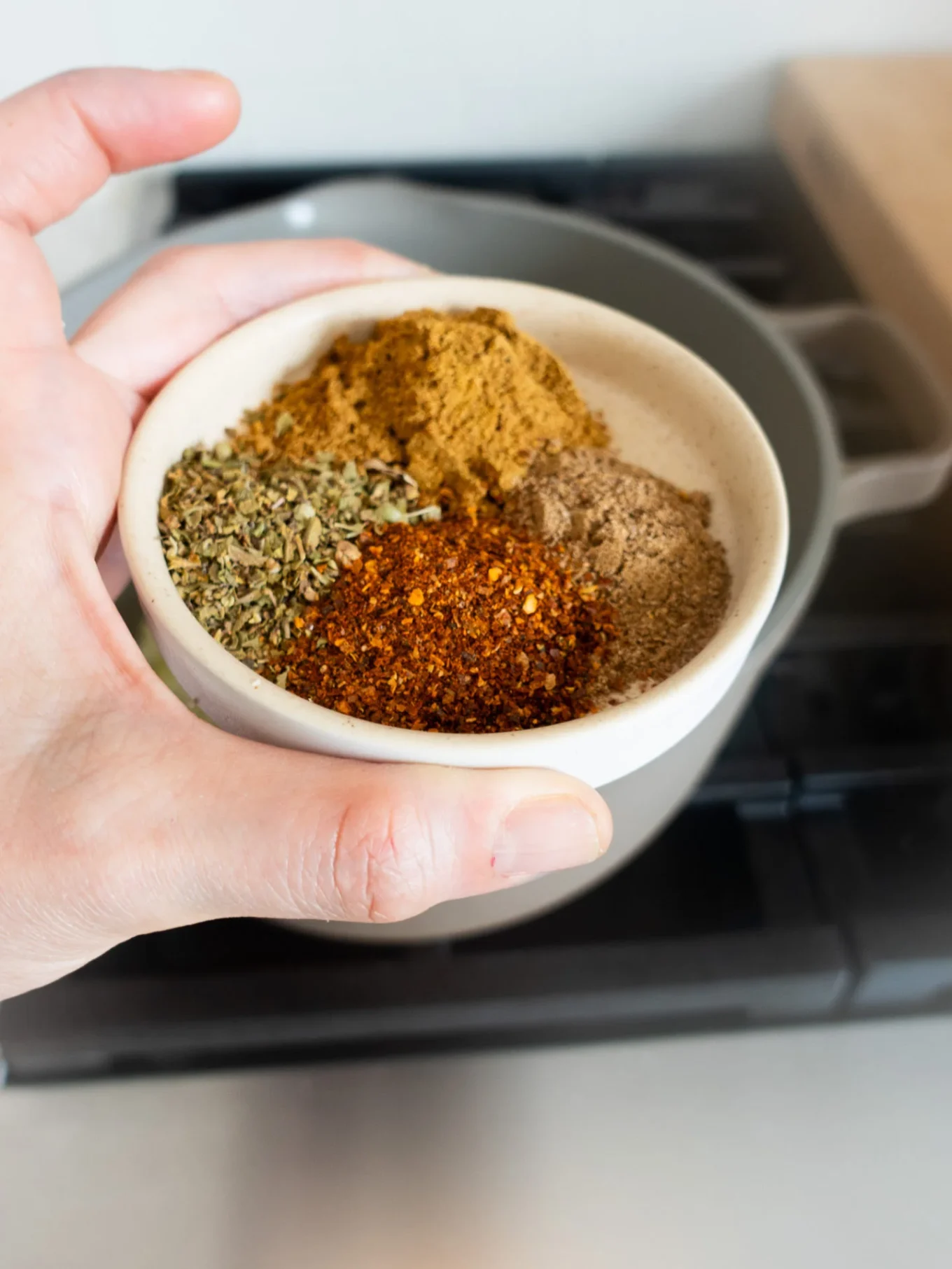 A hand holds a small white bowl filled with four types of ground spices&mdash;brown, green, red, and tan&mdash;perfect for adding bold flavor to creamy White Bean Chili simmering in a pot on the stove. The spices are grouped, showcasing their distinct textures and colors.