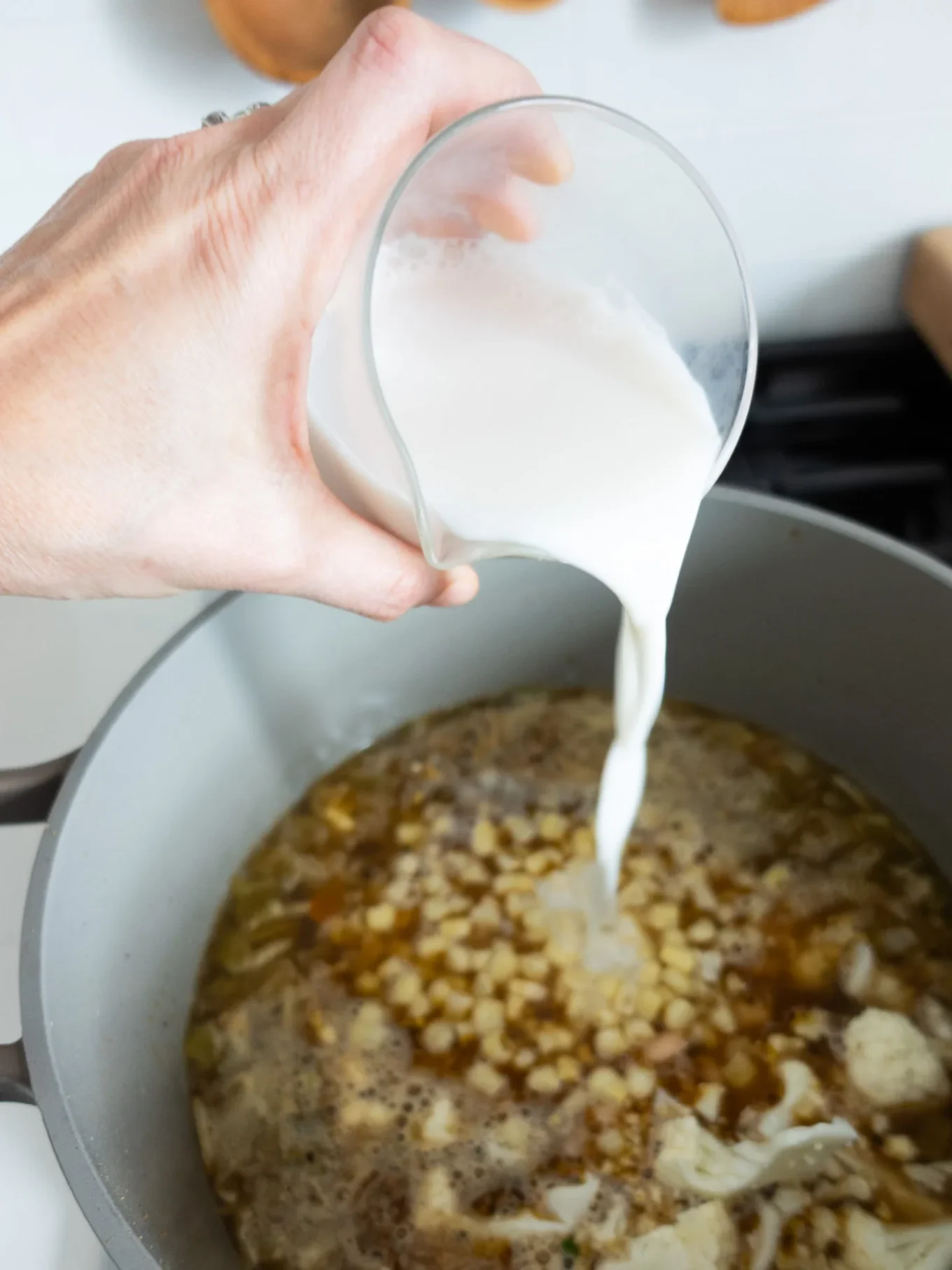 A hand pours milk into a pot of bubbling corn and broth, preparing a creamy soup reminiscent of Creamy Chili or White Bean Chili on the stovetop.