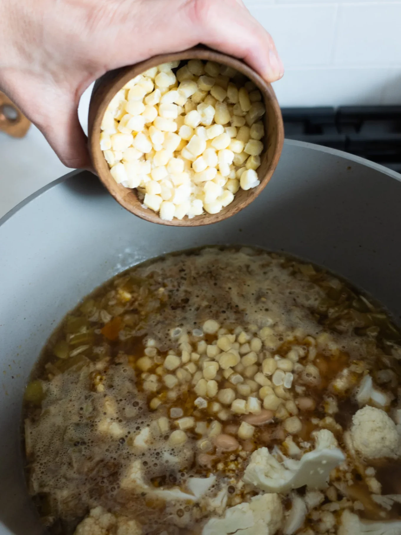 A hand pours a small wooden bowl of white hominy kernels into a simmering pot of creamy cauliflower and white bean chili. The gray pot sits on a stovetop, with a white tiled backsplash in the background.