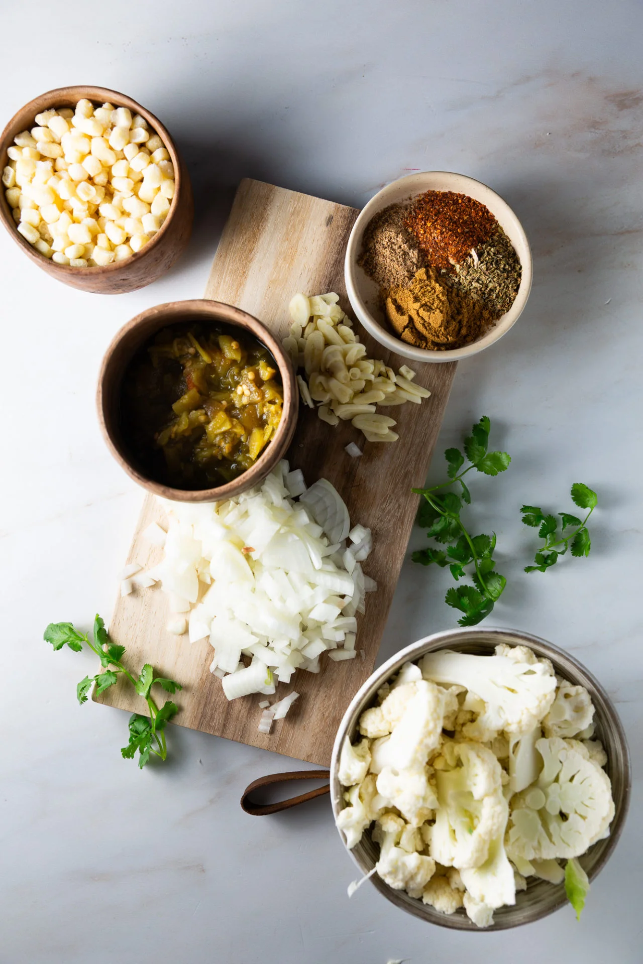 A wooden board with chopped onions and sliced garlic sits on a marble surface. Surrounding it are bowls containing corn, cauliflower florets, green chili sauce, and assorted ground spices&mdash;perfect ingredients for a creamy White Bean Chili. Fresh cilantro leaves are scattered nearby.