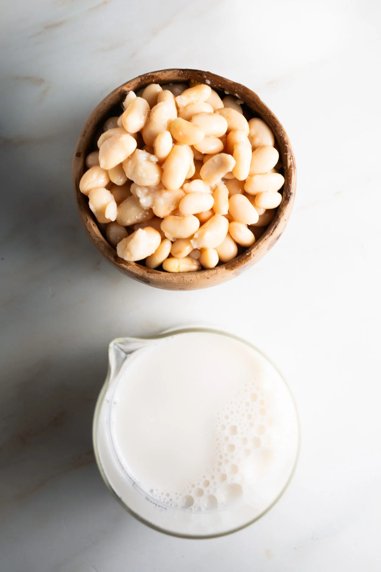 A wooden bowl filled with white beans, perfect for Creamy Chili or White Bean Chili, sits above a glass measuring cup of plant-based milk on a white marble surface. The tidy arrangement is viewed from above.