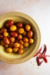 A rustic green bowl holds Spicy Mexican Potatoes—baby potatoes coated in a reddish seasoning. Next to the bowl on a pale surface are whole dried red chilies and scattered chili seeds, hinting at their fiery flavor.