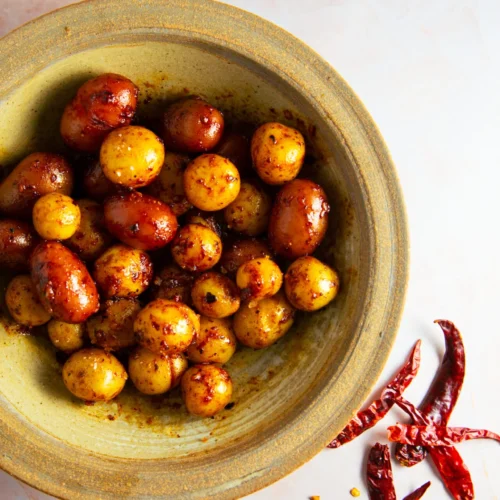 A rustic green bowl holds Spicy Mexican Potatoes—baby potatoes coated in a reddish seasoning. Next to the bowl on a pale surface are whole dried red chilies and scattered chili seeds, hinting at their fiery flavor.