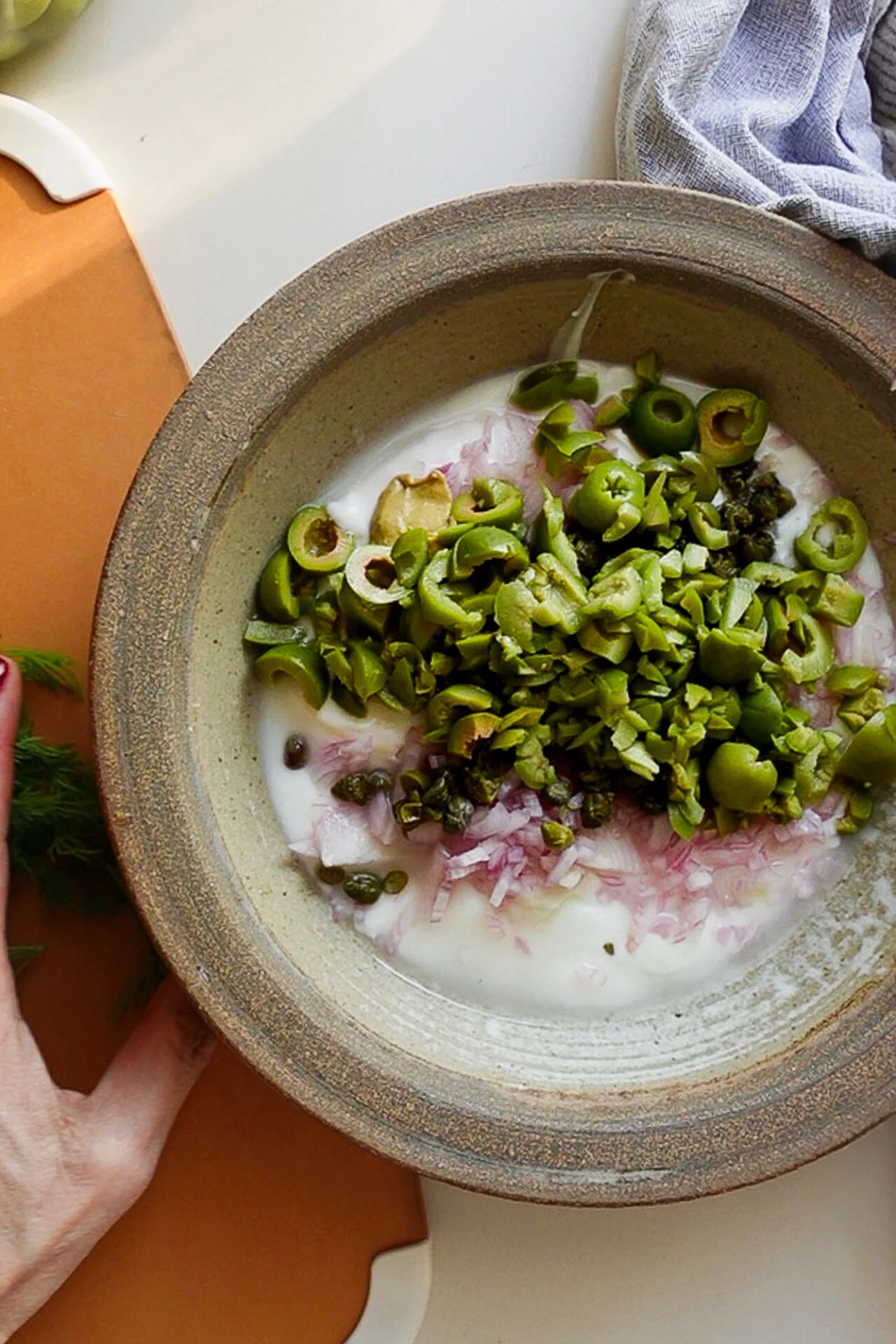 Un bowl de cerámica con yogur, cebolla morada picada, aceitunas verdes rebanadas y alcaparras, listos para preparar el aderezo de la ensalada de papas