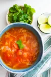 A blue bowl filled with Mexican Cabbage Soup (Caldo de Repollo), topped with a cilantro leaf, sits on a white surface. To the side are a plate of fresh cilantro, lime wedges in a small bowl, and a white and green striped cloth napkin.