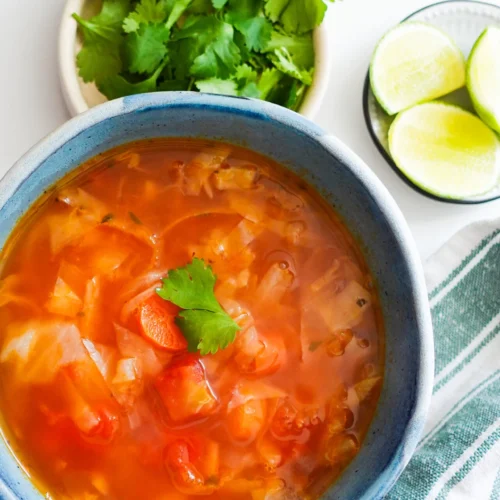 A blue bowl filled with Mexican Cabbage Soup (Caldo de Repollo), topped with a cilantro leaf, sits on a white surface. To the side are a plate of fresh cilantro, lime wedges in a small bowl, and a white and green striped cloth napkin.