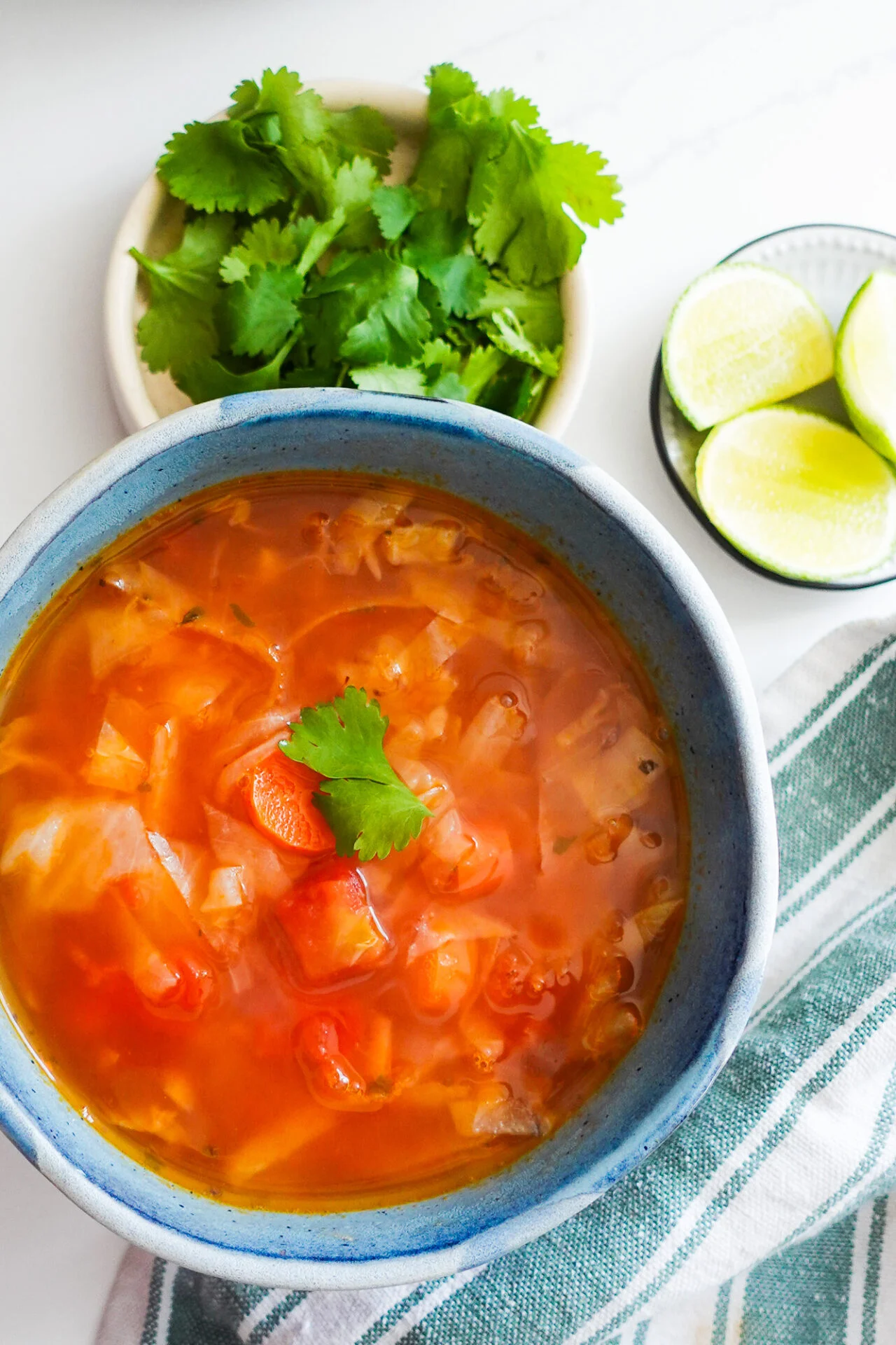 A blue bowl filled with Mexican Cabbage Soup (Caldo de Repollo), topped with a cilantro leaf, sits on a white surface. To the side are a plate of fresh cilantro, lime wedges in a small bowl, and a white and green striped cloth napkin.