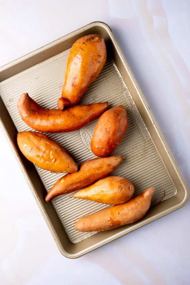 Una bandeja de horno que contiene siete Camotes Horneados enteros, cada uno con la piel dorada y ligeramente brillante. Los camotes var&iacute;an de forma y tama&ntilde;o, y la bandeja descansa sobre una superficie de m&aacute;rmol p&aacute;lido.