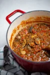 Una olla de hierro fundido roja llena de un guiso de lentejas espeso y sustancioso —que recuerda a un reconfortante ragú vegano— contiene tomates, setas y verduras. La olla descansa sobre una superficie blanca junto a un paño de cocina a rayas, con una cuchara de madera parcialmente visible dentro del guiso.